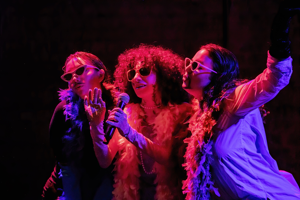 Three actors wearing sunglasses and feather sing into a microphone. They are lit with a red and purple spotlight.